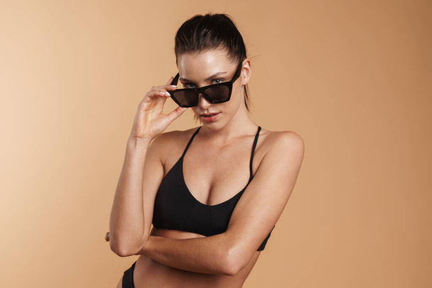 Young brunette woman in swimsuit and sunglasses looking at camera isolated beige background - Φωτογραφία, εικόνα