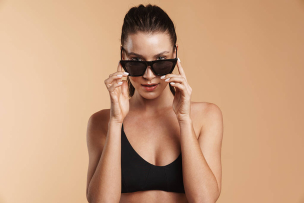 Young brunette woman in swimsuit and sunglasses looking at camera isolated beige background - Фото, изображение