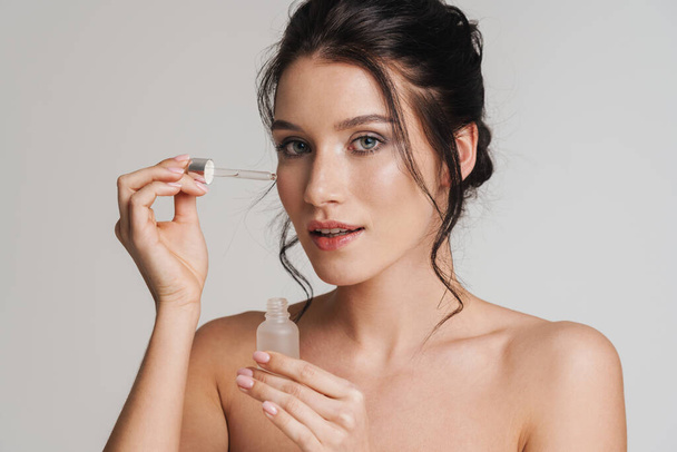 Young white woman with brown hair pulled up applying essential oil on her face with pipette isolated over gray background - Foto, Imagem