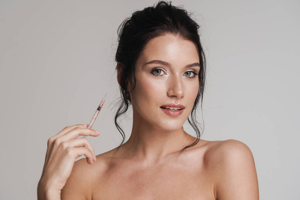 Young healthy white woman with brown hair pulled up holding syringe for aesthetic procedure isolated over gray background - Foto, Imagem