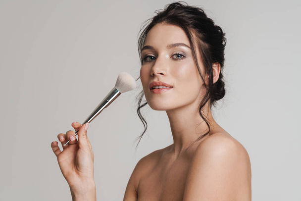 Young white woman with brown hair pulled up holding makeup blush brush isolated over gray background - Fotografie, Obrázek