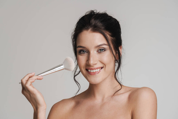 Young smiling white woman with brown hair pulled up holding makeup blush brush isolated over gray background - Foto, afbeelding