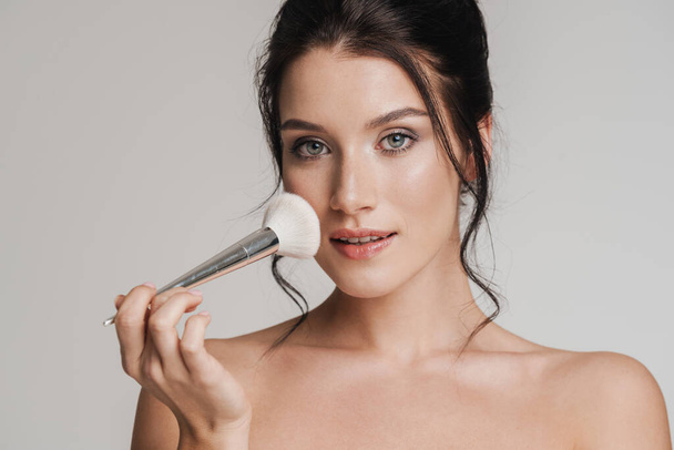 Young white woman with brown hair pulled up holding makeup blush brush isolated over gray background - Fotoğraf, Görsel