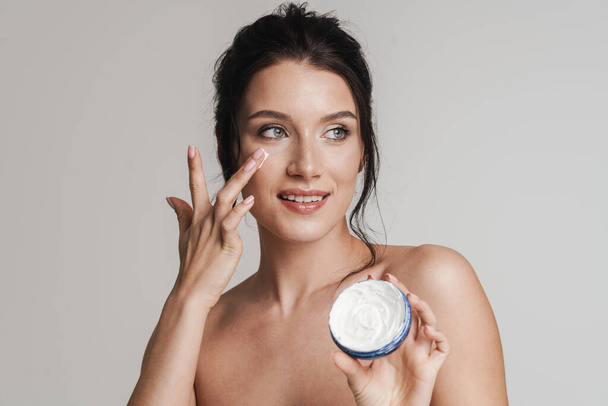 Young smiling white woman with brown hair pulled up applying facial cream from a jar isolated over gray background - 写真・画像