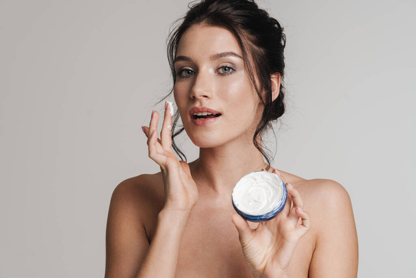Young white woman with brown hair pulled up applying facial cream from a jar isolated over gray background - Foto, Imagem