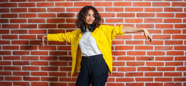 Full length view of the beautiful young sexy girl in a yellow jacket listening to music in headphones on a brick wall background. Excited woman dancing in front of the camera. Positive emotions concept - Fotografie, Obrázek