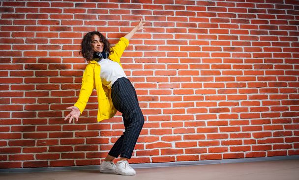 Positive human emotions. Portrait of happy emotional teenage girl with headphones, bending back one hand up. Wearing bright yellow cotton jacket and white shirt. Brick wall background - Фото, изображение