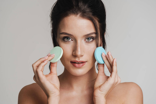 Young white woman with brown hair pulled up applying makeup using beauty blender sponge isolated over gray background - Photo, image