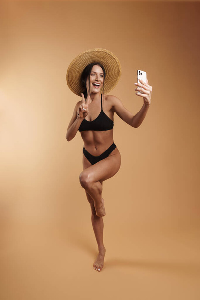 Young woman in swimsuit gesturing while taking selfie on cellphone isolated beige background - Zdjęcie, obraz