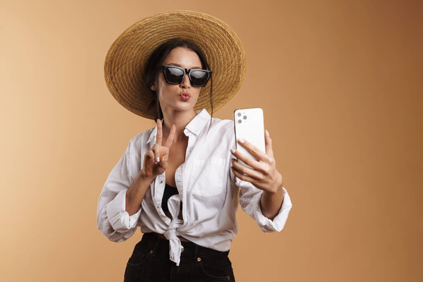 Mujer joven haciendo gestos mientras toma selfie en el teléfono celular aislado sobre fondo beige - Foto, imagen