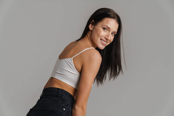 Smiling young white woman with long brunette hair wearing crop top standing posing over gray background side view - 写真・画像