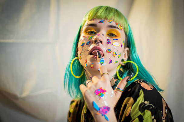 Closeup fashion portrait young pretty beautiful girl with green and blue hair. Beautiful fashion girl with luxury professional makeup and funny emoji stickers glued on the face. Young woman - 写真・画像