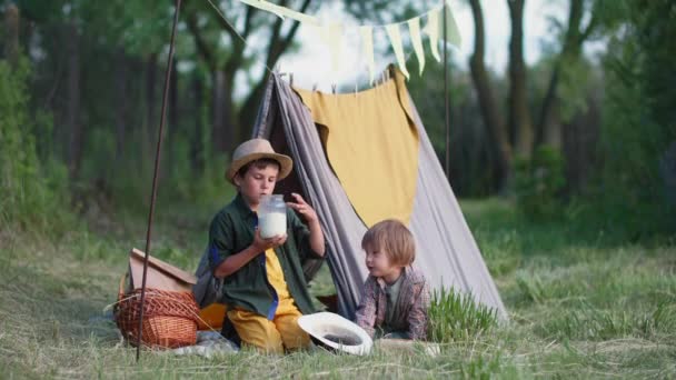 relationship of children, caring boy gives milk from can to his younger brother while relaxing in teepee at picnic among trees - Footage, Video