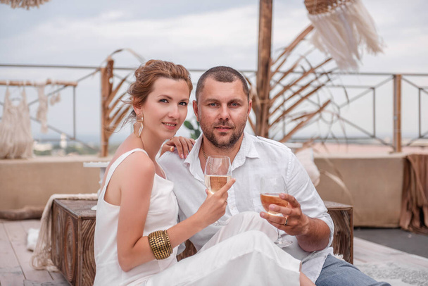 A loving couple sits in a wooden hut in the Moroccan style, drinking white wine. Newlyweds celebrate their anniversary. Young woman laughs, hugs bearded man on open luxury roof terrace with sea view. - Photo, Image