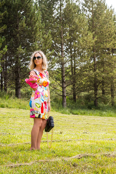 Blond haired model posing in a grove showing off her pink shoes.The girl is wearing a multi-colored dress above the knees.Ideal photography for fashion product.Photo taken in portrait format. - Fotoğraf, Görsel