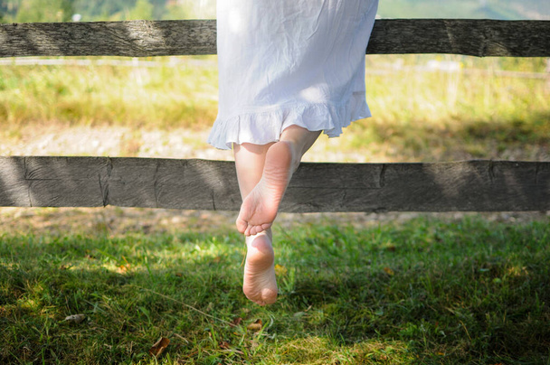 Ξυπόλητη καυστική γυναίκα με λευκό φόρεμα κοντά σε ξύλινο φράχτη. Φυσικό στυλ καλοκαίρι - Φωτογραφία, εικόνα