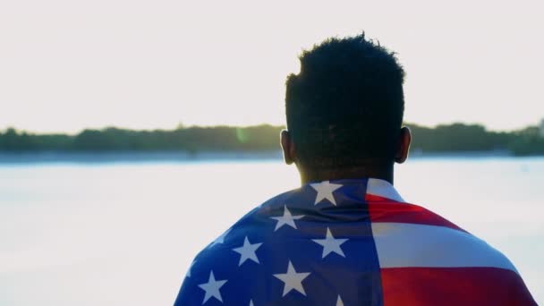 Hombre afroamericano con bandera americana sobre hombros mira a la distancia al amanecer - Metraje, vídeo
