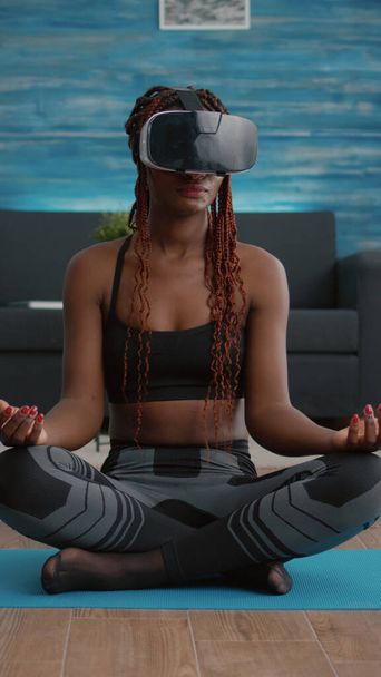 Fit black woman wearing virtual reality headset while sitting on yoga map - 写真・画像