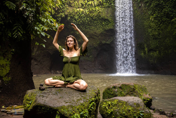 posa loto yoga. Donna caucasica seduta sulla pietra, meditando, praticando pranayama. Mani in alto. Esercizio di respirazione. Occhi chiusi. Flusso di energia. Ritiro di yoga. Cascata Tibumana, Bali - Foto, immagini