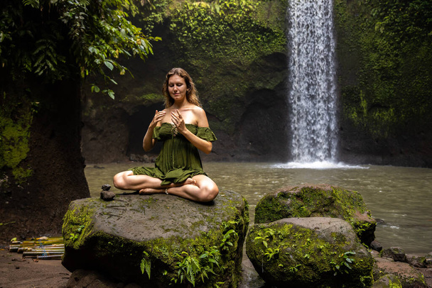 Йога-лотос пози. Кавказька жінка сидить на камені, медитуючи, практикуючи йогу і пранаяму. Вправи для дихання. Заплющені очі. Потік енергії. Відступ йоги. Водоспад Тібумана, Балі (Індонезія). - Фото, зображення