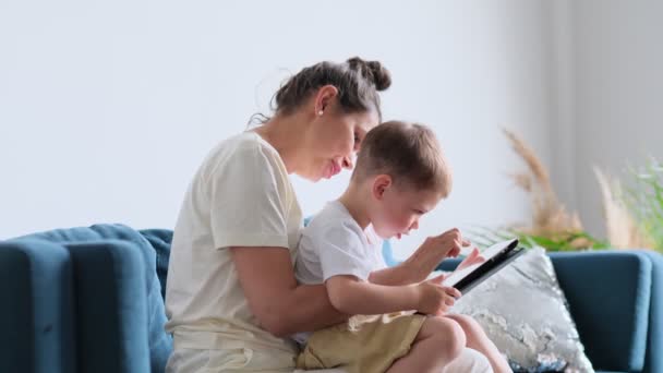 madre e hijo con la tableta en casa. Madre mostrando contenido multimedia en línea a su hijo en una tableta sentada en un sofá en la sala de estar en el interior de una casa - Metraje, vídeo