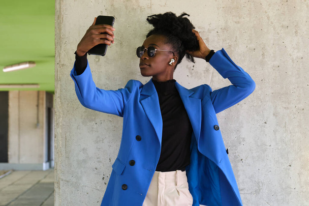 Portrait of young african woman wearing wireless earbuds, taking a selfie with her smartphone, wearing blue jacket suit and sunglasses over a concrete wall. - Valokuva, kuva