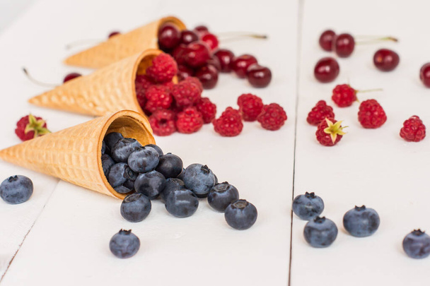 appetizing waffle cones with blueberries, raspberries, cherries on a white wooden background. Summer dessert. - Foto, imagen