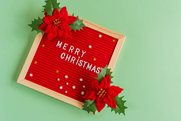 Merry christmas greeting postcard in minimalism style. letter board with greetings and red piosentia flowers on green background. - Photo, image