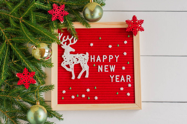 English text happy new year and toy deer on a felt board in a wooden frame. letterboard on white wooden background with fir tree and gold baubles. - Photo, image