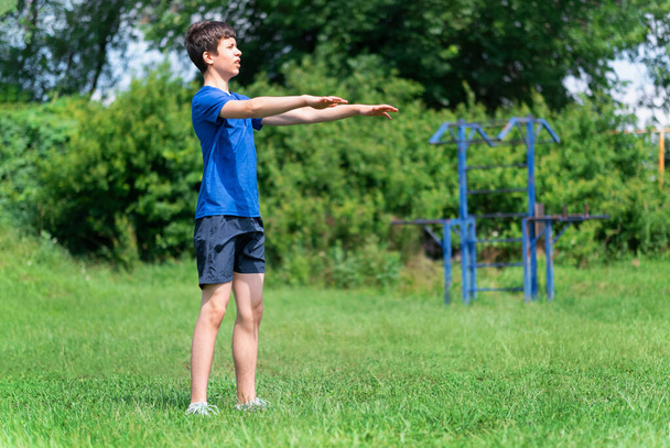 мальчик-подросток, занимающийся на открытом воздухе, спортивной площадкой во дворе, садится на корточки и ведет разминку, здоровый образ жизни - Фото, изображение