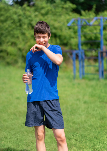 teenage boy exercising outdoors, sports ground in the yard, he opens a bottle of water and drinks, healthy lifestyle - Photo, Image