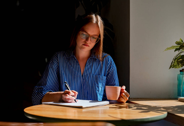 Young woman writing plans in notebook, sitting at wood desk in cafe with coffee cup and sunlight - Фото, изображение