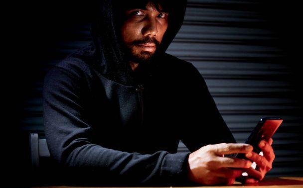 Mysterious man in hoodie on dark background. Dangerous criminal. Silhouette of man in the hood, dark mysterious man hoodie, murderer, hacker, anonymus on the black background with free space - Photo, Image