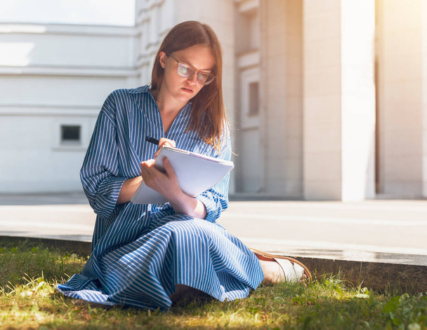 Female student sitting on grass near university and learning, writing smth, reading, holding textbook. Young smart woman in glasses studying alone outdoors in summer - 写真・画像