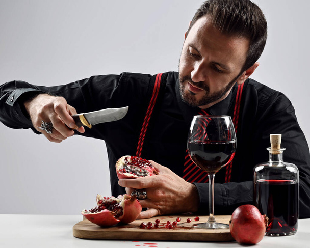 Brutal bearded man in black shirt with striped tie and suspenders sits at table eating fresh pomegranate and juice - Foto, imagen