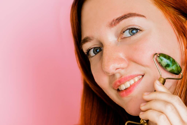 A beautiful redhead girl massages her face with a jade roller. The girl smiles on a pink background. - Photo, Image