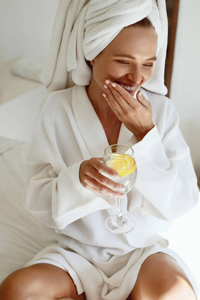 Laughing european girl drink lemonade after shower. Young smiling beautiful woman wear bathrobe and wrapped bath towel on head. Concept of rest with spa treatment. - Photo, image