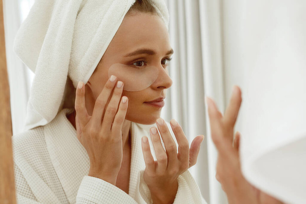 European girl applying under eye patch on face and watch in mirror. Young concentrated beautiful woman wear bathrobe and wrapped bath towel on head. Concept of face skin care - Φωτογραφία, εικόνα