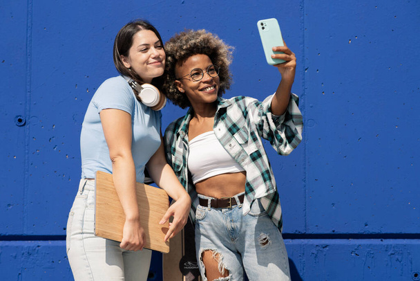 dois amigos multi-étnicos tomando selfie, com fones de ouvido e laptop, em uma parede azul - Foto, Imagem