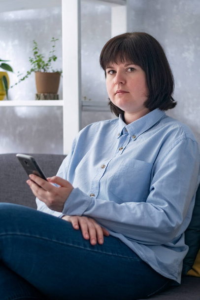 Poważna kobieta siedzi na kanapie z telefonem w rękach. Smm manager pracujący w domu. - Zdjęcie, obraz