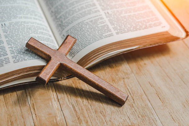 Крест поверх открытой библии на деревянном столе с оконным светом. - Фото, изображение