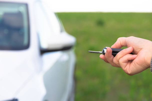 De hand van een vrouw houdt de sleutel van een nieuwe witte auto vast en opent deze op een zonnige zomerdag tegen een achtergrond van groen gras. Selectieve focus. Close-up - Foto, afbeelding