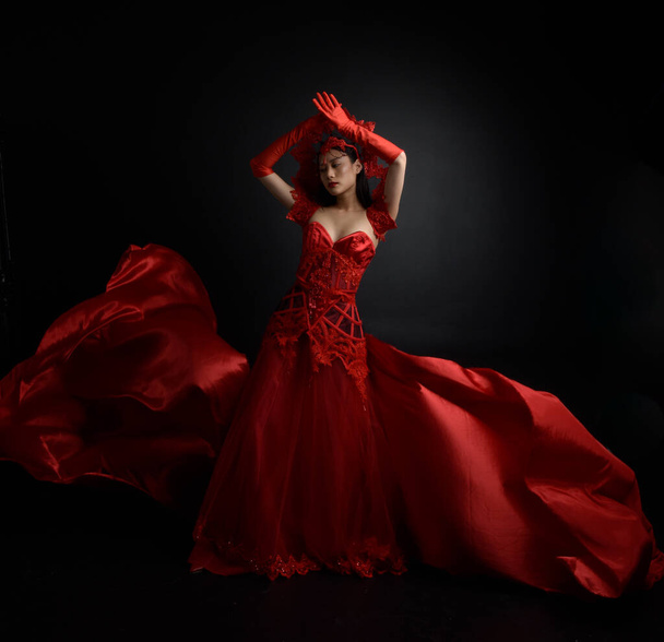 dramatic portrait of beautiful young asian woman wearing red corset, long opera gloves and ornate gothic queen crown. Graceful posing isolated on a dark studio background. - Foto, Imagem