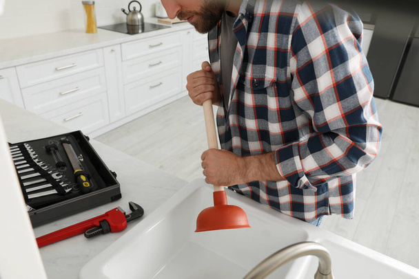 Young man using plunger to unclog sink drain in kitchen, closeup - Foto, afbeelding