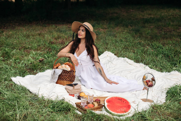 Portret van een jong mooi meisje met zelfs witte tanden, een mooie glimlach in een strohoed en lange witte jurk picknicken in de tuin - Foto, afbeelding
