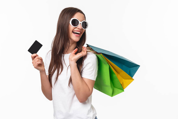 Shopping, leisure and lifestyle concept. Portrait of carefree happy young woman release stress with buying new things, paying with credit card, hold shop bags on shoulder and looking back smiling - Фото, изображение