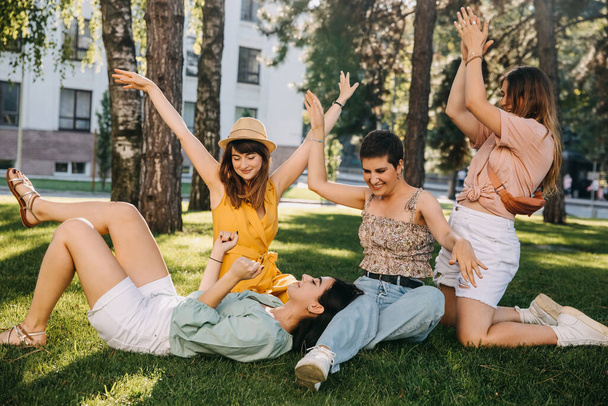 Gruppe von vier Freundinnen in einem Park, Spaß haben, auf grünem Gras sitzen, tanzen. - Foto, Bild