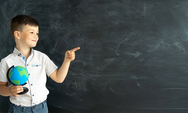 a smiling schoolboy at school with a globe stands in front of a chalk board and points his finger at a place for text. Education and study. board in the background. back to school. - Photo, Image