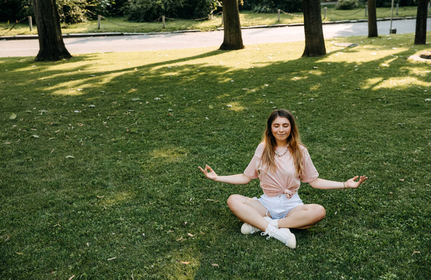 Молода жінка сидить на зеленій траві в парку в позі йоги лотоса
. - Фото, зображення