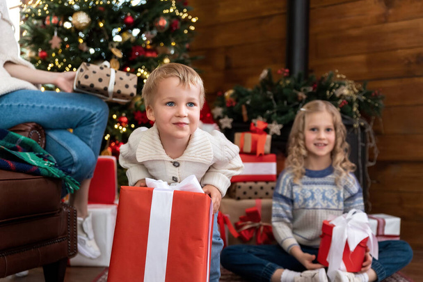 Retrato lindo divertido niño emocionado bebé con la hermana y la madre abren gran caja de regalo blanco regalo de Navidad contra el árbol de Navidad decorado en casa luz de la mañana. Feliz concepto de vacaciones de infancia - Foto, imagen